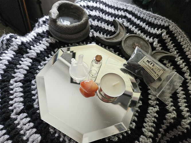 A mirror containing spell items including a small funnel, a red-orange carnelian crystal, a tealight candle in a metal holder, a corked glass vial, and a labeled plastic bag of “Modern Magick Witches Brew #5,” all arranged on a black and white crocheted surface.