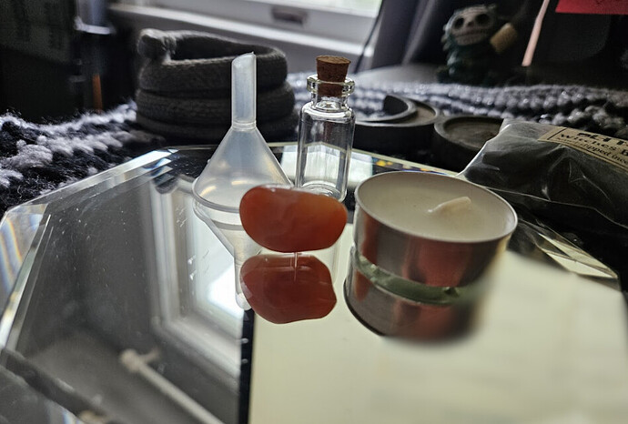A mirror containing spell items including a small glass bottle with cork, a tea light candle in a metal holder, and a red-orange carnelian crystal, on a black and silver altar cloth with other spiritual items.