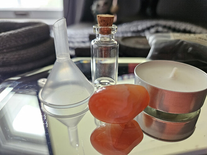 A close-up view of a mirror containing spell items including a small funnel, a red-orange carnelian crystal, a tealight candle in a metal holder, and a corked glass vial, on a black and silver altar cloth.